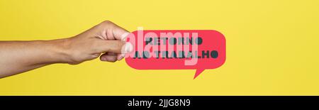 the hand of a man holding a red paper sign with the text back to work written in portuguese, on a yellow background, in a panoramic format to use as w Stock Photo