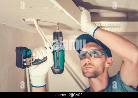 Closeup of Caucasian Contractor in His 40s Focused on Installation of the Gypsum Plasterboard Using Electric Screwdriver. Construction Site Industrial Stock Photo