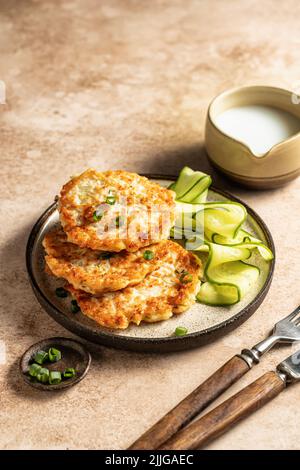 Vegetarian zucchini fritters with green onion and cucumber served with cream sauce on brown background with cutlery. Text space, vertical orientation Stock Photo