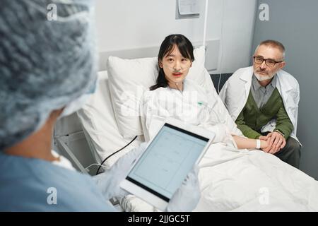 Unrecognizable doctor holding digital tablet talking to young Asian patient and her father in emergency room Stock Photo