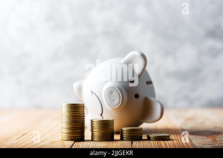 A sad piggy bank behind stacks of euro coins symbolizing the fall of monetary assets. White moneybox with coins graph. Crisis concept Stock Photo