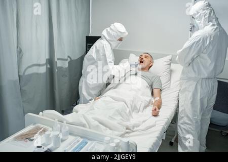Unrecognizable doctors wearing white protective suits with masks testing mature man for infectious or bacterial disease Stock Photo