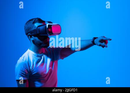 Man in white t-shirt wearing VR goggles using metaverse technology innovation in the virtual world. AR Concept over neon lighting and dark background Stock Photo