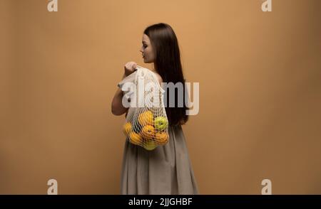 Back of woman in beige dress holding string shopping reusable mesh bag with fruits on a shoulder. Zero waste concept. Space for text. Reusable eco Stock Photo