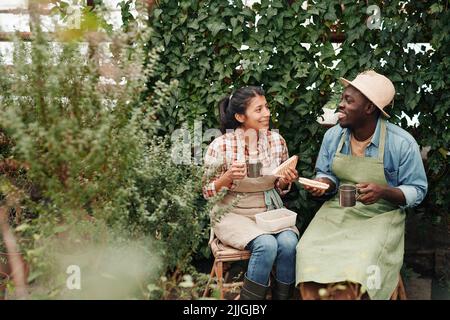 Two young ethnically diverse co-workers wearing aprons enjoying having lunch together in modern greenhouse Stock Photo