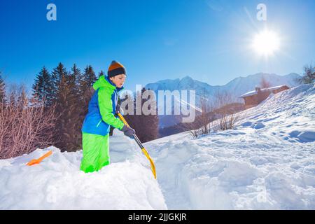 Boy in ski outfit build snow fortress with shovel over mountains Stock Photo