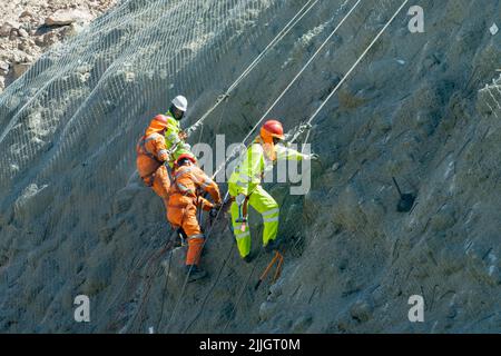 Construction workers on ropes set rebar anchors on wire netting to protect cars from falling rocks in a canyon in Chile. Stock Photo