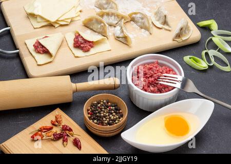Meat filling in bowl, allspice, egg in bowl. Raw gyoza dumplings and stuffed dough pieces on cutting board. Top view. Black background Stock Photo
