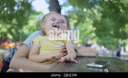 Closeup of cute little girl plays with mom while sitting in the park. Close-up of mother and daughter playing while sitting on a park bench in a stree Stock Photo