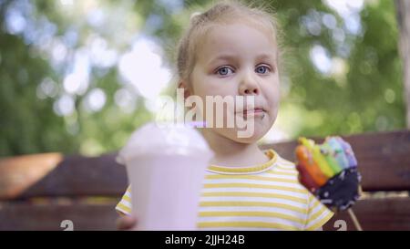 ittle girl eats a colorful gingerbread and holds a milkshake in her hand. Close-up of cute child girl sitting on park bench and eating cookies with a Stock Photo