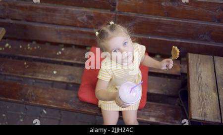 ittle girl eats a colorful gingerbread and holds a milkshake in her hand. Close-up of cute child girl sitting on park bench and eating cookies with a Stock Photo