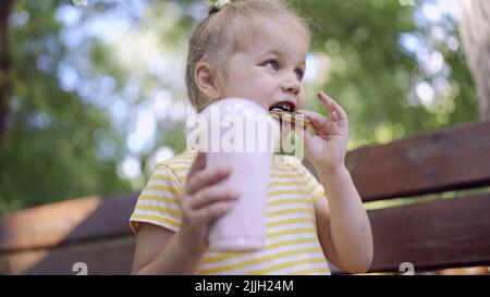 ittle girl eats a colorful gingerbread and holds a milkshake in her hand. Close-up of cute child girl sitting on park bench and eating cookies with a Stock Photo