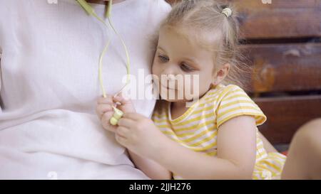 Close-up of cute little girl is sitting next to mom and playing with her headphones. Close-up of child girl sitting on park bench and examining mother Stock Photo
