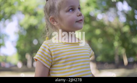 Cute little girl listens to music and sings along. Close-up portrait of child girl stands in a city park and listens to music Stock Photo