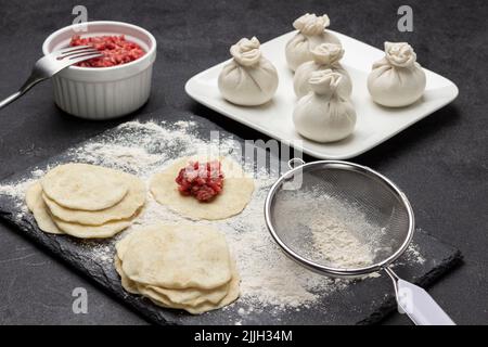 Pieces of dough and meat on dough, flour in sieve on stone board. Raw khinkali dumplings on white plate. Minced meat and fork in bowl. Top view. Black Stock Photo