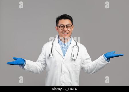 Smiling adult japanese male doctor in white coat, glasses and protective gloves hold empty space on hands Stock Photo