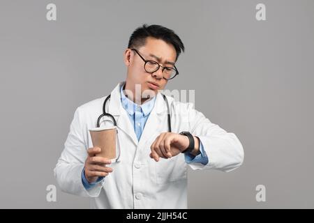 Serious busy middle aged japanese male doctor in white coat and glasses with cup of drink takeaway Stock Photo