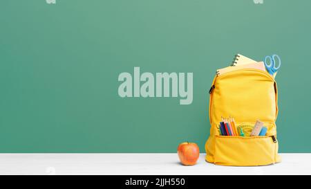 Backpack with different colorful stationery and apple on table and green background. Banner design back to school concept background. Stock Photo
