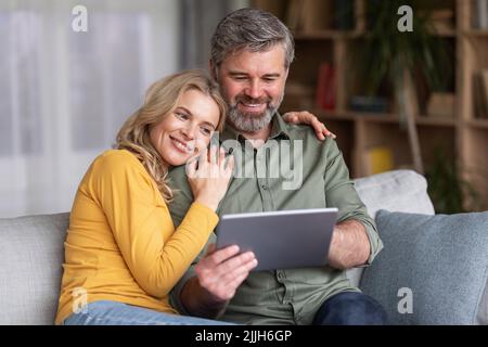 Great App. Happy Middle Aged Spouses Using Digital Tablet Together At Home Stock Photo
