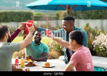 Cheerful multiracial male friends with food on table toasting drinks while having fun in yard Stock Photo