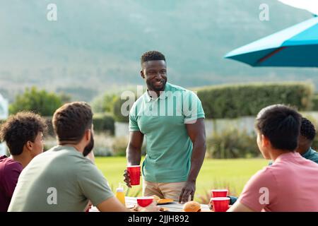 Happy multiracial young man standing and talking with friends while enjoying drinks and food in yard Stock Photo