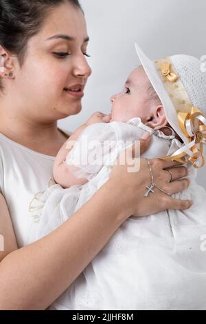 beautiful young latin mother, holding her baby with a silver chain entangled in her hand and with a cross pendant. baby baptism in catholic church. co Stock Photo
