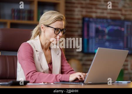 Portrait of adult businesswoman working with laptop at office. Female trader texting messages, doing paperwork with stock market graphs on background Stock Photo
