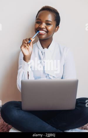 Portrait of an african-american woman sitting on the floor with laptop, smiling while looking at camera. Online education, remote work. Beautiful Stock Photo