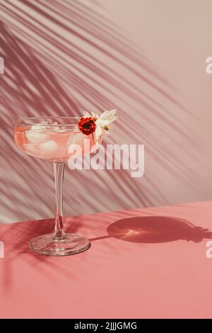 Summer cocktail with pink champagne with ice on the table with shadows from the sun. On a pink wall background with palm leaves, front view. Stock Photo