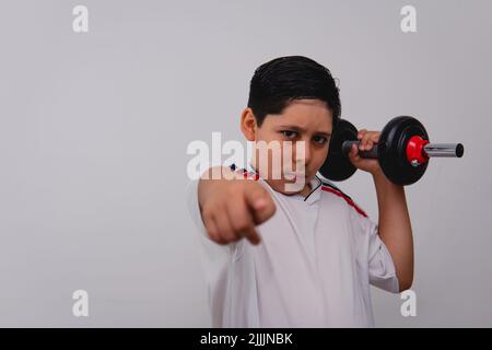 A boy dressed in white with a sports shirt carrying a black dumbbell pointing at the camera. A sporty boy using gym weights with a challenging look. Stock Photo