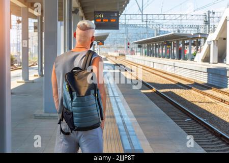 Lonely young man in a t shirt and a backpack on his back stands with his back to the camera on the railway platform waiting for an electric train. Stock Photo