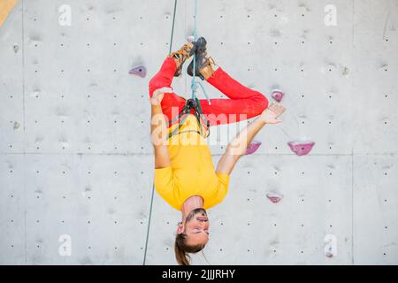 A man hangs upside down in a lotus position on a climbing wall. The man is wearing a yellow T-shirt and red pants. Stock Photo