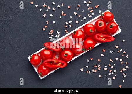 Fresh washed cherry tomatoes and paprika slices on a white oblong dish with pink salt on a black concrete table Stock Photo