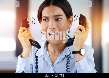 I cant do everything at once. a young call centre agent sitting alone in the office and feeling overwhelmed while answering two phone calls. Stock Photo