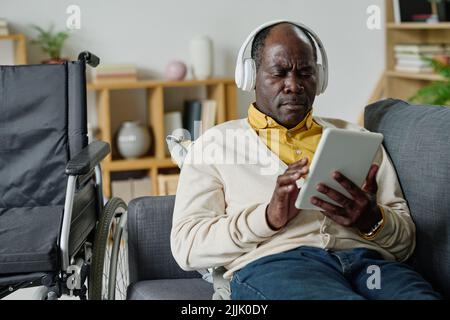 African mature man with disability using digital tablet for musical therapy for relaxation on sofa in living room Stock Photo