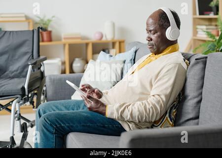 African man with disability sitting on sofa in wireless headphones and using tablet pc for listening music in the room Stock Photo