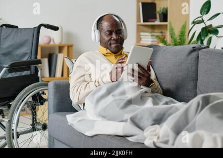 African mature man with disability lying on sofa under the blanket with digital tablet and using headphones for listening to music Stock Photo