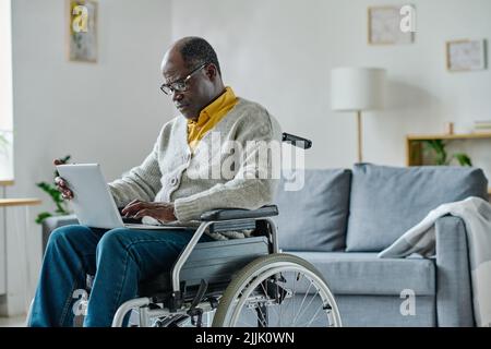 African mature man with disability sitting in wheelchair and communicating online using his laptop at home Stock Photo