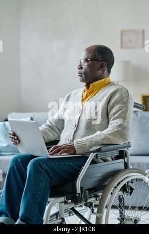 African mature man sitting on wheelchair looking away with pensive expression while working online using laptop at home Stock Photo