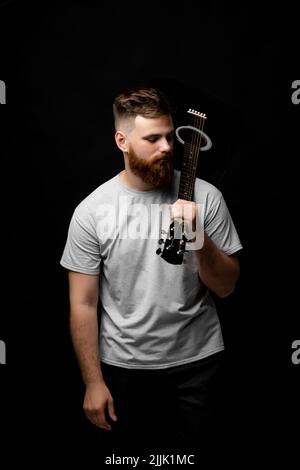 Brutal handsome bearded man in grey t-shirt with a guitar on a shoulder. Portrait of a male singer and guitarist. Stock Photo