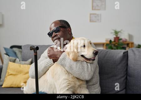 African senior man in glasses with bad sight embracing his individual guide dog while sitting on sofa at home Stock Photo