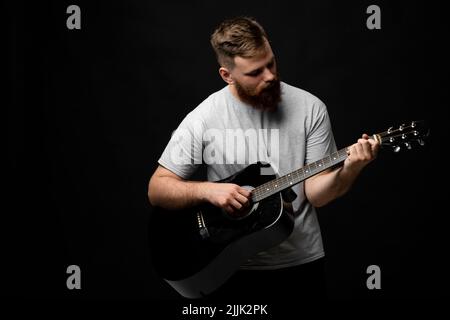 Concentrated brutal handsome bearded man in grey t-shirt playing acoustic guitar. Portrait of a male singer and guitarist. Stock Photo