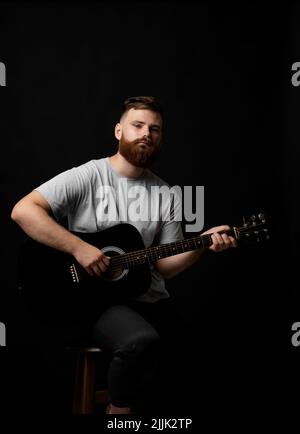 Bearded brutal guitarist plays an acoustic guitar in a black room and looking in the camera. The concept of music recording, rehearsal or live Stock Photo