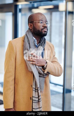 African confident businessman in elegant coat putting on the scarf around his neck, he leaving the office building Stock Photo