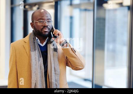 African businessman in stylish coat talking on mobile phone while walking along the office corridor after work Stock Photo