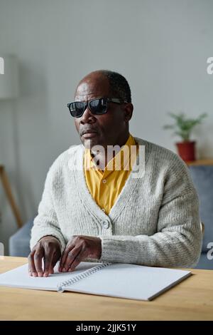 African American blind man in dark glasses, sitting at table and reading book in braille language Stock Photo