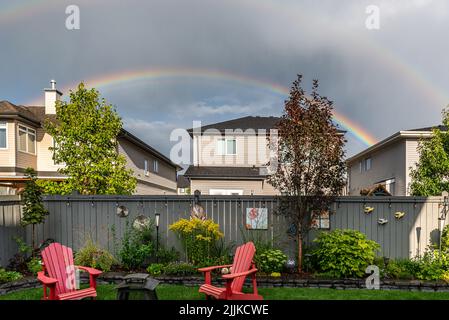 Rainbow over houses in a neighborhood and pink chairs in a yard of a house Stock Photo