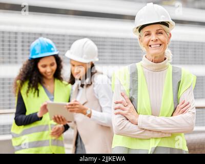 Were building the future. Cropped portrait of an attractive mature female engineer standing with her arms folded with her colleagues in the background Stock Photo
