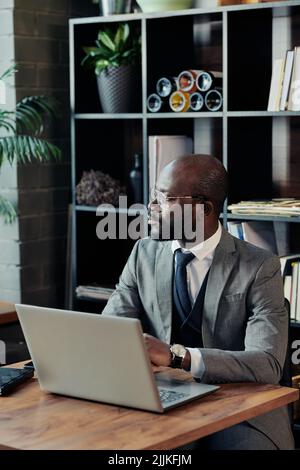 African man in suit looking through the window with pensive sight while working online on laptop in cafe Stock Photo