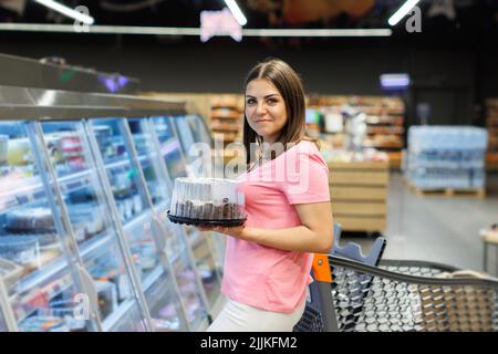 A beautiful smiling woman stands in a large grocery store near a grocery cart and chooses a holiday cake. A woman is holding a box of cake while stand Stock Photo